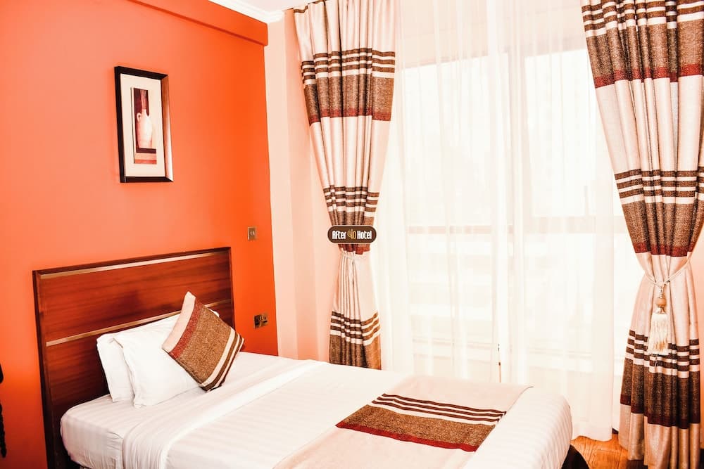 A cozy hotel room features an inviting bed with striped bedding, vibrant orange walls, patterned curtains, and soft natural light, ideal for travelers seeking comfort and relaxation.