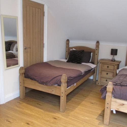 A cozy attic bedroom with wooden flooring, rustic wooden beds adorned with brown bedding, a small bedside table with a lamp, and a mirror reflecting the serene ambiance, ideal for travelers seeking a quaint and comfortable stay.