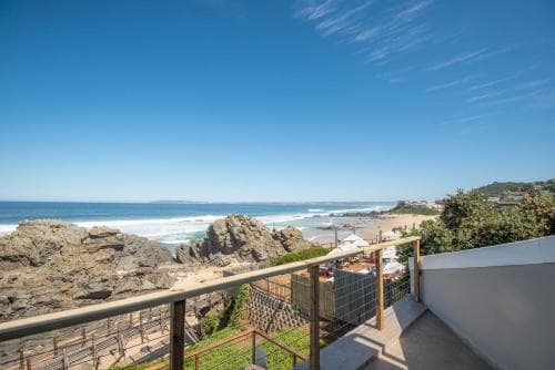 A serene coastal balcony overlooks a rocky beach with turquoise waters under a clear blue sky, ideal for travelers seeking relaxation and scenic views.