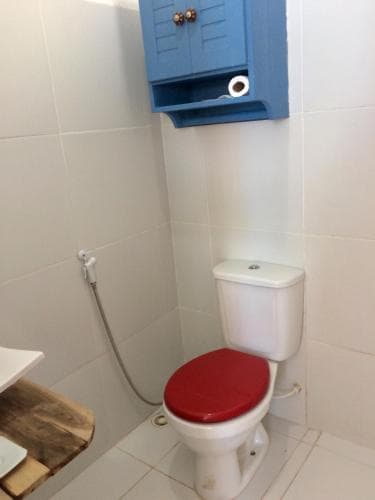 A compact bathroom with white tiled walls features a toilet with a red seat, a blue cabinet above, and a wooden bench, ideal for travelers seeking efficient space utilization in accommodations.