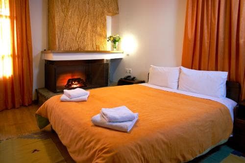 A cozy hotel room features a warmly lit fireplace, orange curtains, neatly folded towels on the bed, and a vase of flowers, ideal for travelers seeking comfort and relaxation.