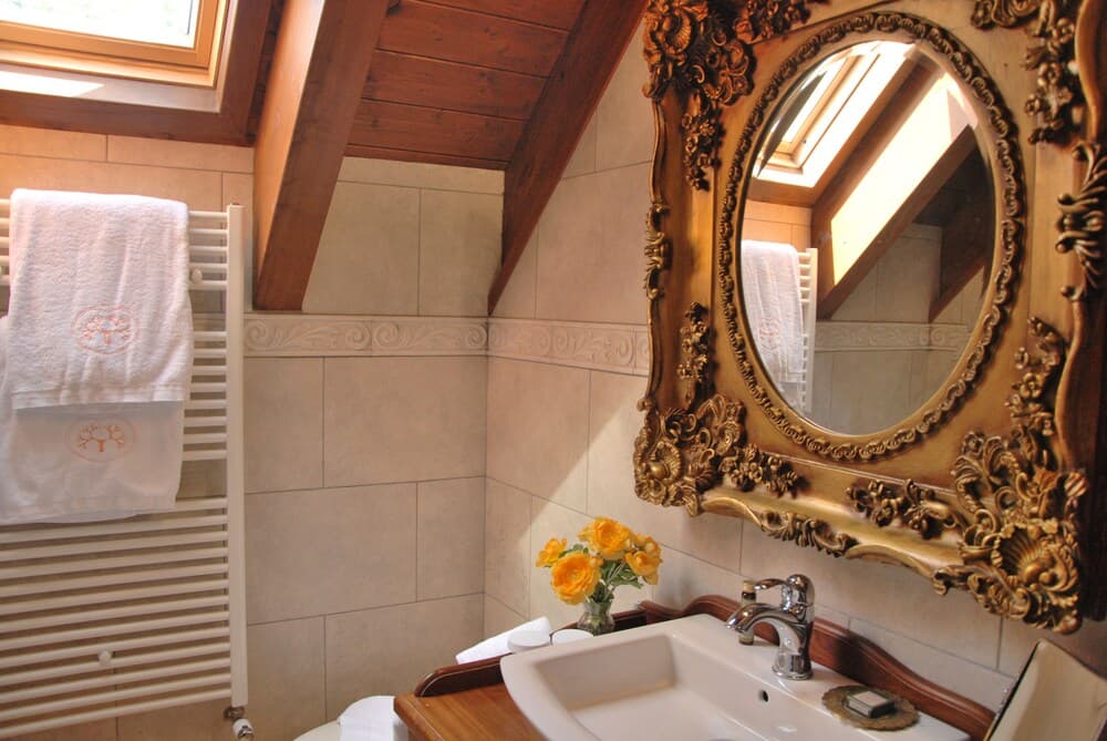 A cozy attic bathroom features a luxurious gold-framed mirror, tiled walls, a wooden beam ceiling, and a vase of yellow flowers, ideal for travelers seeking a charming and elegant accommodation experience.