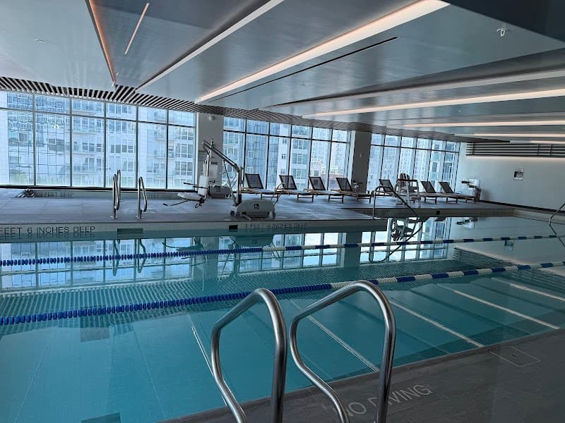 A serene indoor swimming pool at the Renaissance New York Flushing Hotel features modern lounge chairs, large windows with urban views, and sleek design elements, ideal for travelers seeking relaxation and fitness during their stay.
