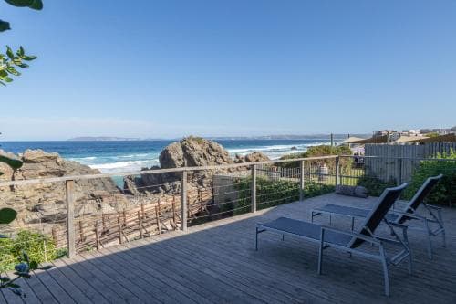 A serene coastal deck offers breathtaking ocean views with lounge chairs, wooden railings, and rocky shores, perfect for a relaxing seaside getaway.