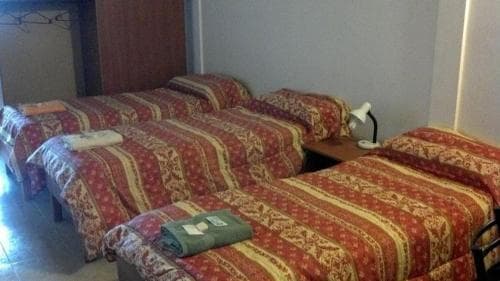 A cozy budget hotel room features three neatly arranged beds with patterned red and gold bedding, a small nightstand with a lamp, and a towel on the bed, ideal for travelers seeking affordable accommodations.