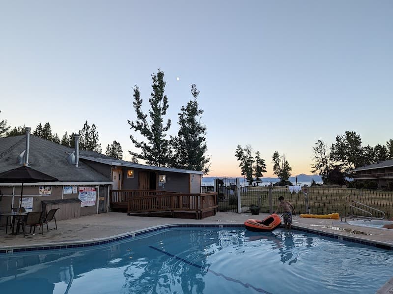 A serene evening at a lakeside resort features a calm pool area with a child playing near an inflatable raft, surrounded by tall pine trees and a cozy cabin, ideal for family vacations seeking relaxation and natural beauty.