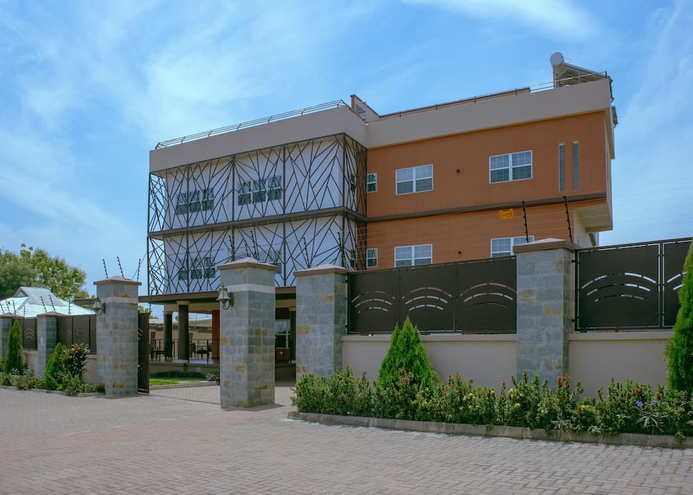 A modern residential building with a striking geometric facade stands behind an elegant stone gate adorned with lush greenery, ideal for travelers seeking upscale accommodation in a serene suburban setting.