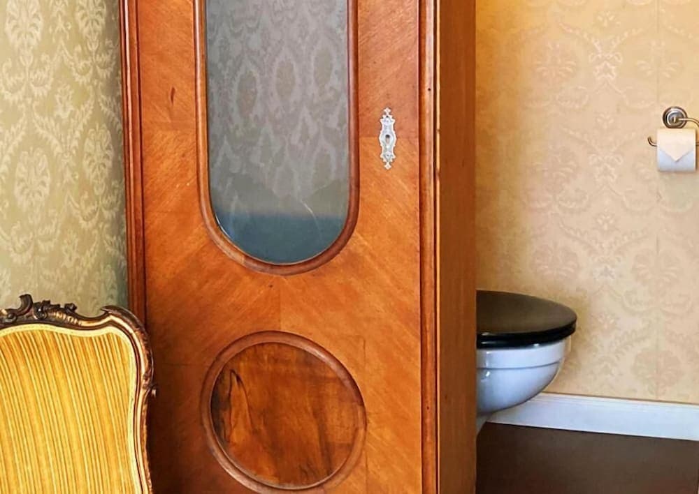 A quaint bathroom with vintage wallpaper exudes charm, featuring an ornate wooden cabinet with oval cutouts, a classic toilet, and a decorative chair, ideal for travelers seeking unique accommodations with historical ambiance.