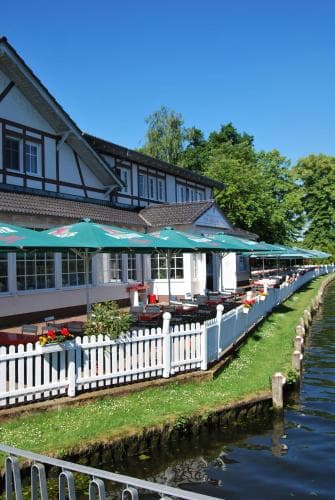 A serene riverside café with green umbrellas and white picket fences invites travelers to relax and enjoy waterfront dining under a clear blue sky.