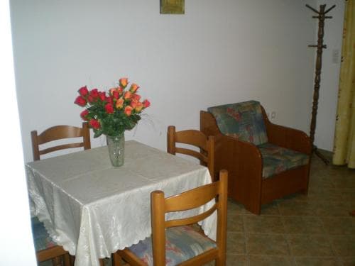 A cozy dining nook features a wooden table draped in a white cloth, adorned with vibrant red and orange flowers, set against a minimalist backdrop with a cushioned armchair and rustic coat rack, ideal for travelers seeking a warm and inviting accommodation space.