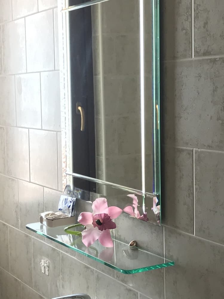 A serene bathroom scene features a sleek glass shelf holding a delicate pink flower and toiletries, set against tiled walls, ideal for travelers seeking modern and minimalist accommodations.