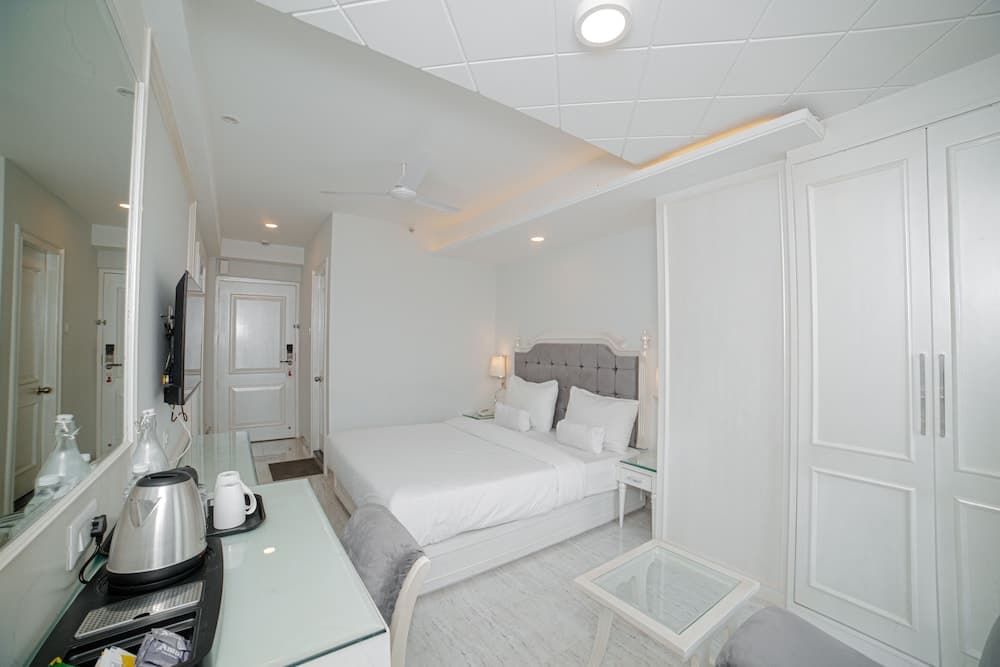 A serene hotel room features a cozy bed with white linens, sleek white furniture, a glass-top desk with a kettle, and soft lighting, ideal for travelers seeking comfort and relaxation.