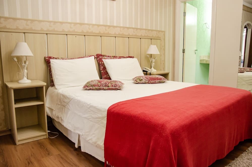 A cozy hotel room features a neatly made bed with white linens and a red throw blanket, flanked by wooden nightstands with lamps, set against striped wallpaper, ideal for travelers seeking comfort and relaxation.