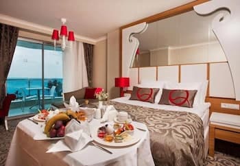 A cozy hotel room with a scenic ocean view through large glass doors, featuring a neatly arranged bed, vibrant red accents, a breakfast spread on a table, and a modern chandelier, ideal for travelers seeking comfort and relaxation by the sea.