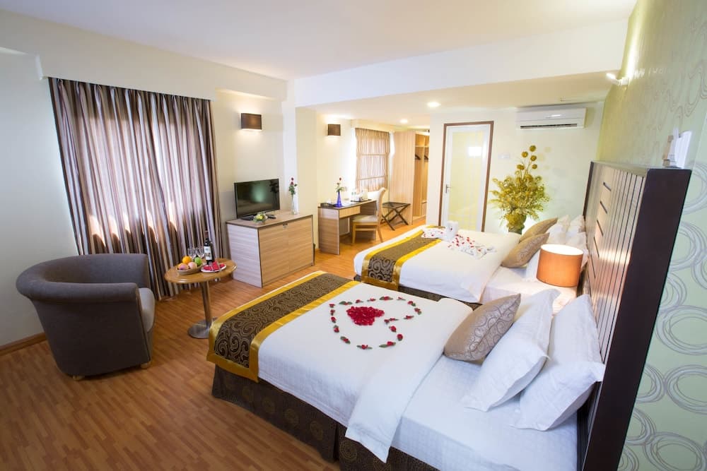 A cozy hotel room with two neatly arranged beds adorned with a rose petal heart, warm lighting, wooden furnishings, and a serene ambiance ideal for romantic getaways or comfortable stays.