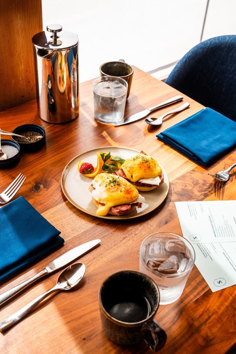 A cozy dining table at Stephanie Inn & Dining Room in Cannon Beach, Oregon, features a delicious Eggs Benedict plate, elegant blue napkins, and a French press, perfect for a relaxing brunch experience.