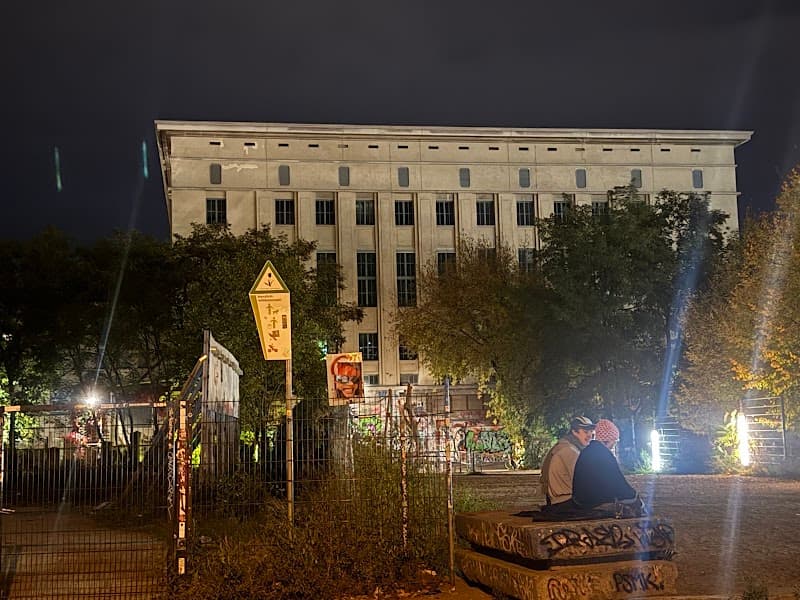 A serene night scene outside the iconic Berghain-Panorama Bar in Berlin, featuring a couple sitting on a graffiti-covered bench under illuminated trees, evoking a contemplative mood ideal for urban nightlife enthusiasts exploring the city's vibrant club culture.