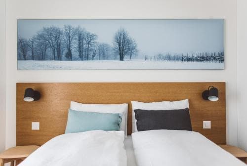 A serene hotel room features a cozy bed with white linens under a wooden headboard, flanked by modern wall sconces, set against a tranquil winter landscape painting, ideal for travelers seeking relaxation.