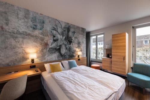 A cozy hotel room features a large bed with white linens, a vibrant floral mural wall, wooden furnishings, and a bright window view, ideal for travelers seeking comfort and modern aesthetics.