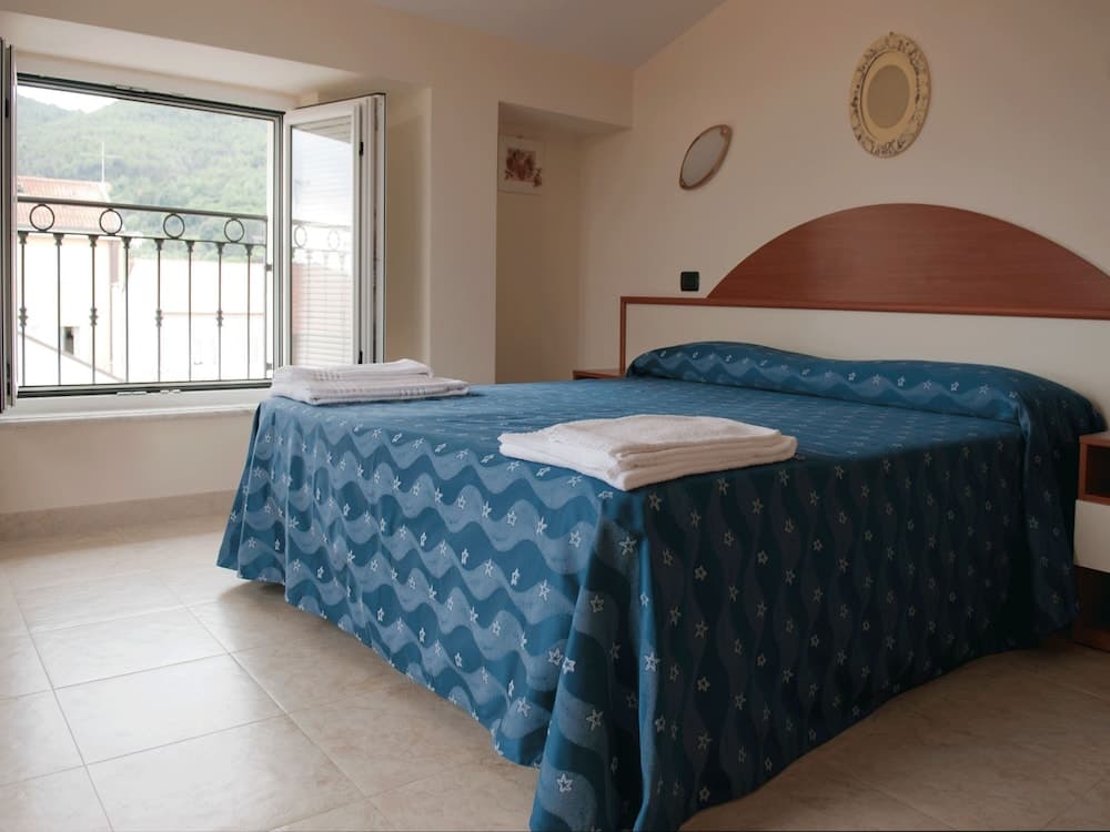A cozy hotel room with a neatly made bed adorned in blue star-patterned bedding, a balcony offering a serene view of green hills, and simple decor, ideal for travelers seeking relaxation and comfort.