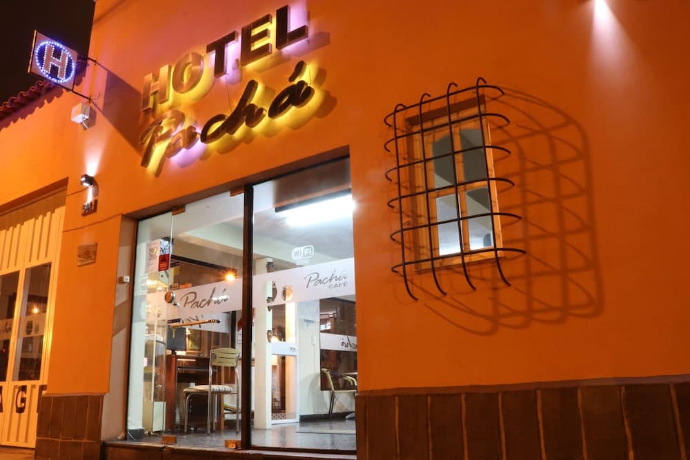 A warmly lit hotel entrance with a vibrant orange facade, featuring a neon "Hotel Pachá" sign and a cozy café interior, creates an inviting atmosphere ideal for travelers seeking a charming urban retreat.
