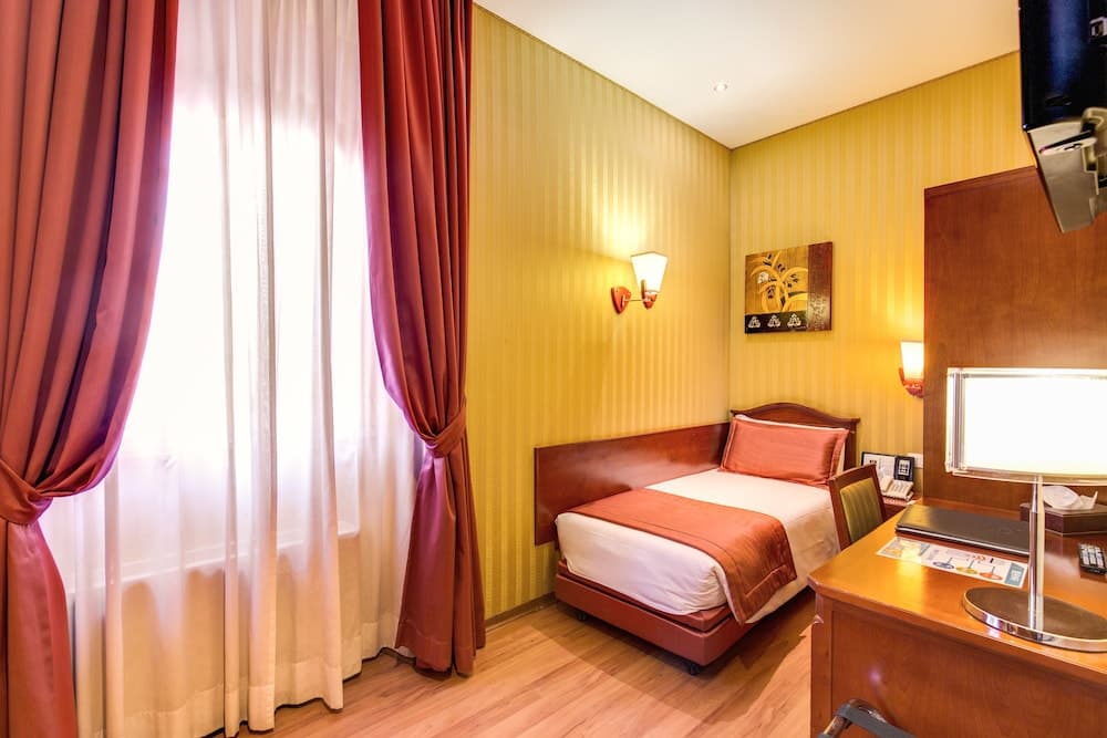 A cozy hotel room with warm yellow walls, a neatly made bed adorned with red accents, a wooden desk with a lamp, and large curtains framing a bright window, ideal for travelers seeking comfort and relaxation.