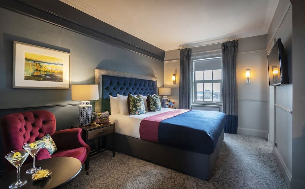 A cozy hotel room features a plush bed with blue and pink bedding, a red velvet armchair, patterned curtains, and a window view, creating an inviting atmosphere ideal for travelers seeking comfort and relaxation.