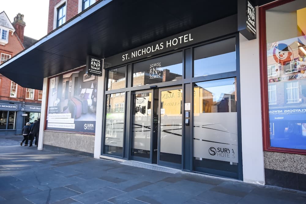 The inviting entrance of St. Nicholas Hotel features modern glass doors and a sleek black awning, set against a charming urban backdrop with brick buildings, ideal for travelers seeking a cozy stay in a bustling city center.