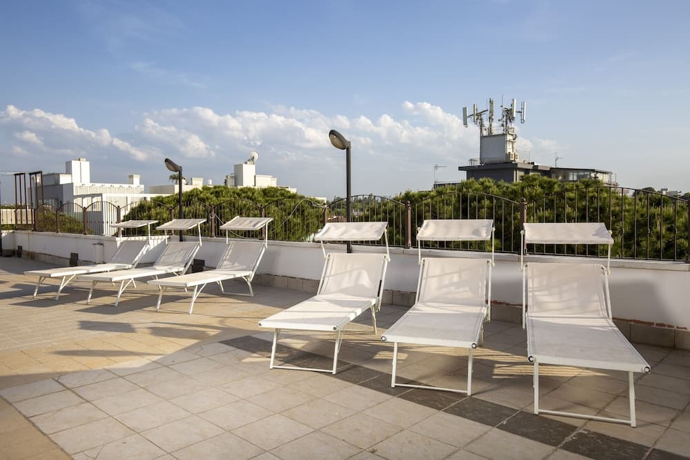 A serene rooftop terrace features white lounge chairs under a clear blue sky, ideal for relaxation and urban sightseeing with a backdrop of greenery and distant buildings.