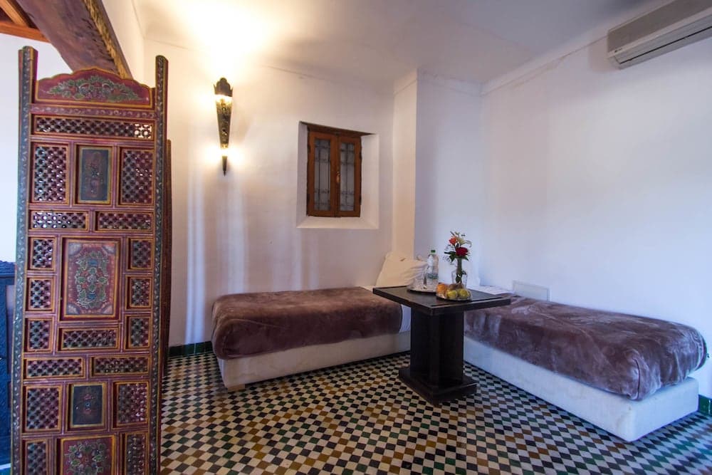A cozy, minimalist guest room with patterned tiled floors, ornate wooden decor, and simple furnishings offers a serene retreat for travelers seeking cultural immersion.