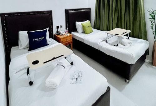 A cozy hotel room features two neatly arranged single beds with white linens, black headboards, and personal trays, set against a backdrop of green curtains and a wooden nightstand, ideal for travelers seeking comfort and convenience.