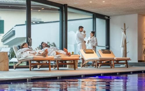Relaxing by the indoor poolside, guests in white robes lounge on wooden chaise lounges under soft lighting, creating a serene spa atmosphere ideal for wellness retreats.