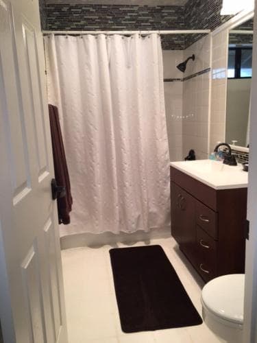 A cozy bathroom with white tiled floors, a black rug, a wooden vanity, and a shower with a white curtain creates a serene atmosphere, ideal for travelers seeking comfort in accommodations.