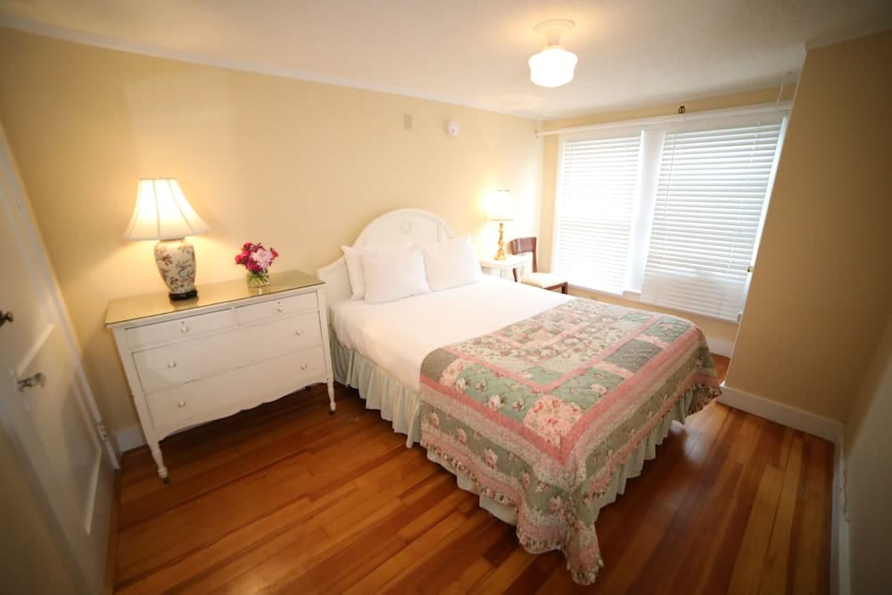 A cozy bedroom with warm yellow walls, wooden floors, a neatly made bed adorned with a floral quilt, and soft lighting from lamps creates an inviting atmosphere ideal for travelers seeking comfort in a vacation rental.