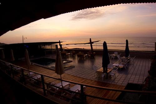 A serene beachside terrace at sunset features lounge chairs, closed umbrellas, and a tiled patio, ideal for relaxing coastal getaways.