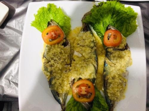 A beautifully plated dish of garlic-covered fish garnished with lettuce and tomato slices on a white plate, perfect for a culinary travel blog showcasing local seafood delicacies.