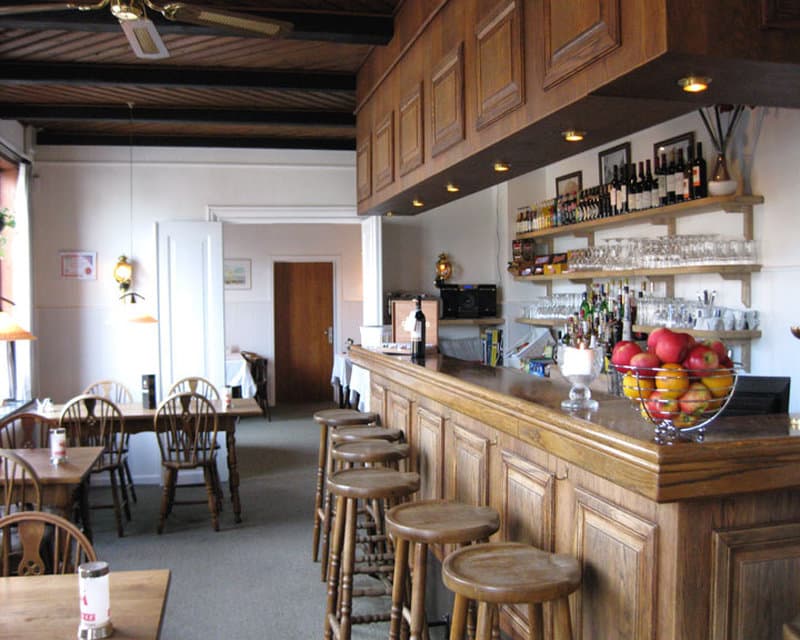 A cozy pub interior features wooden barstools, a well-stocked bar with fruit bowls, and rustic tables set for dining, ideal for travelers seeking a warm and inviting atmosphere for relaxation or socializing.