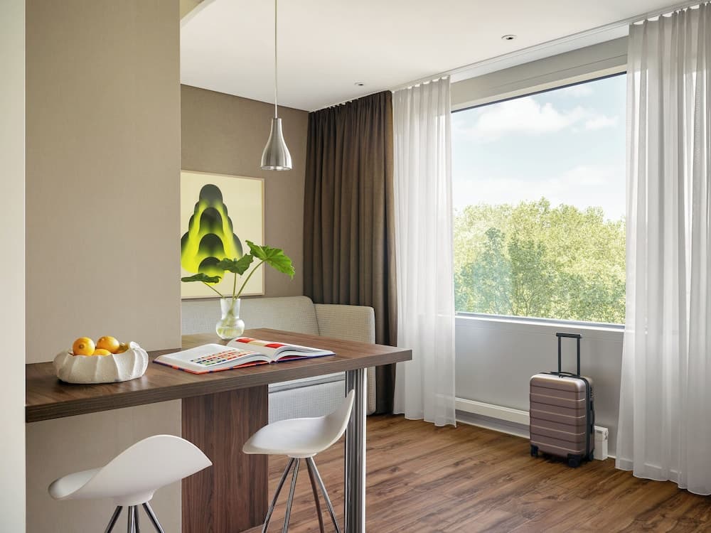 A cozy hotel room features a modern dining area with a wooden table, white chairs, a vibrant painting, and a suitcase by the window, offering a serene view of lush greenery, ideal for travelers seeking comfort and relaxation.