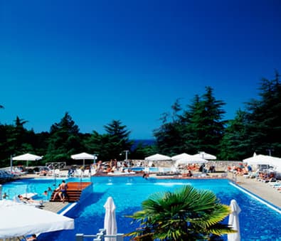A vibrant resort pool area under a clear blue sky, featuring lounge chairs, white umbrellas, and lush greenery, evokes a relaxing vacation atmosphere ideal for travelers seeking leisure and sunbathing spots.