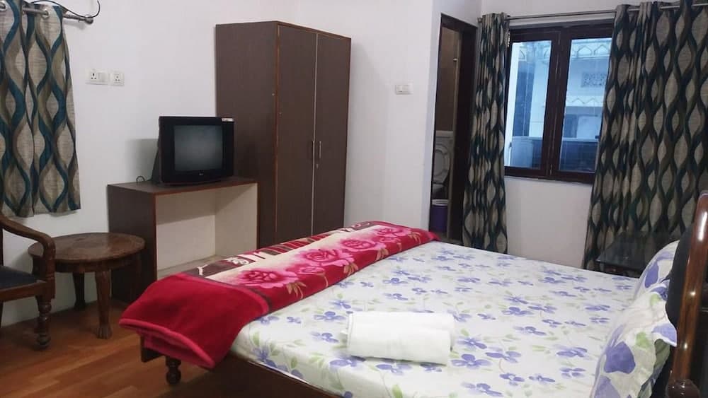 A cozy budget-friendly hotel room features a neatly made bed with floral bedding, a wooden wardrobe, a small TV on a cabinet, patterned curtains, and a calm atmosphere ideal for travelers seeking simple accommodations.