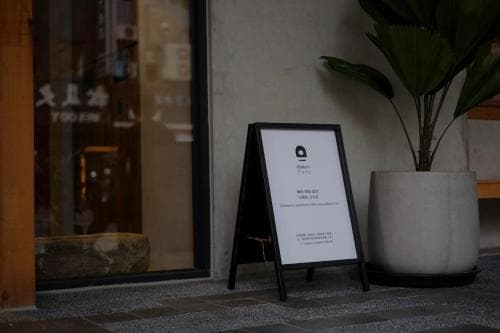 A cozy urban café entrance features a minimalist blackboard sign and a large potted plant, inviting travelers to explore its serene ambiance and modern decor.