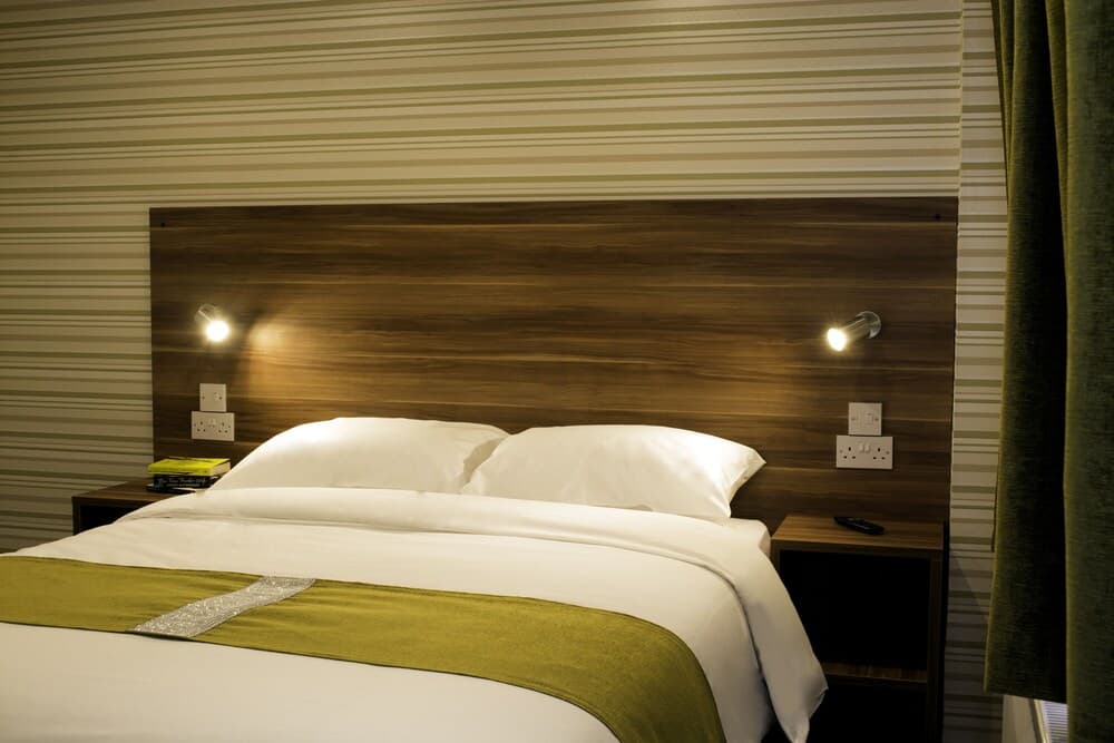 A cozy hotel room features a neatly made bed with white linens and a green throw, flanked by wooden bedside tables with modern lighting, ideal for travelers seeking comfort and style.