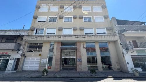A sunny urban street scene features a multi-story beige building with a modern entrance, flanked by small potted plants, ideal for travelers seeking accommodations in a bustling city environment.