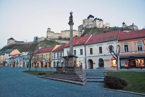 A serene town square features a historic column statue surrounded by cobblestone streets and charming buildings, with a majestic castle perched on the hill in the background, ideal for cultural exploration and photography.