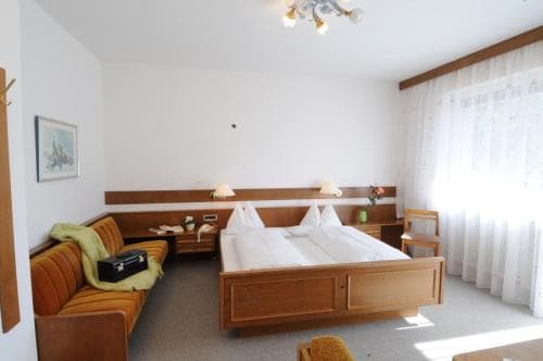 A cozy hotel room features a neatly made wooden bed, a comfortable sofa with a green blanket, soft natural light filtering through sheer curtains, and a calm, inviting atmosphere ideal for travelers seeking relaxation.