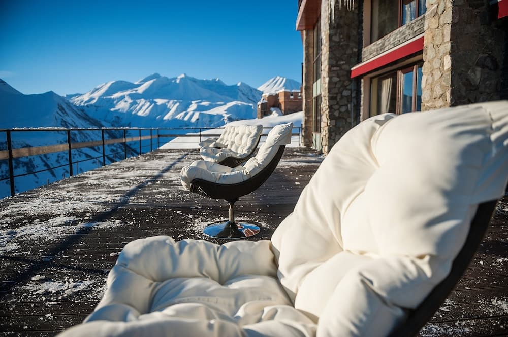 A cozy outdoor seating area on a snowy deck offers breathtaking views of snow-capped mountains, perfect for a serene winter getaway at a mountain lodge.