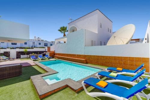 A vibrant resort scene features a sparkling pool surrounded by blue lounge chairs on artificial grass, set against white buildings under a clear sky, ideal for a relaxing vacation getaway.
