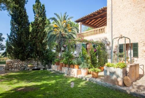 A serene Mediterranean courtyard features lush greenery, potted plants, and a rustic stone house under a clear blue sky, ideal for a tranquil vacation retreat.