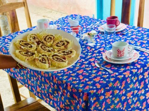 A cozy outdoor dining setup features a vibrant floral tablecloth adorned with freshly baked cinnamon rolls, cups of tea, and glasses, perfect for a relaxing breakfast or brunch experience.