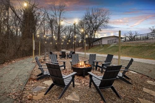 A cozy outdoor gathering spot features Adirondack chairs arranged around a warm fire pit under string lights, set against a tranquil park backdrop at dusk, ideal for a relaxing retreat or group outing.